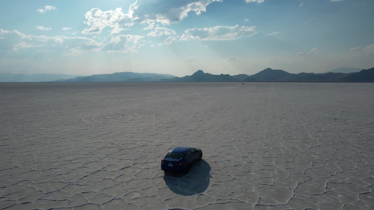 보네빌 소금 평원을 가로지르는 자동차는 극적인 구름 사이로 비치는 햇빛 광선과 배경, 유타, 공중 추적 샷에서 지평선을 만지는 산을 통해 빛나는 독특한 자연적 특징을 가지고 있습니다.
