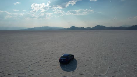 Car-riding-across-Bonneville-Salt-Flats-unique-natural-features-with-beam-of-sunlight-rays-shining-through-dramatic-clouds-and-mountain-touching-horizon-in-background,-Utah,-Aerial-Tracking-Shot