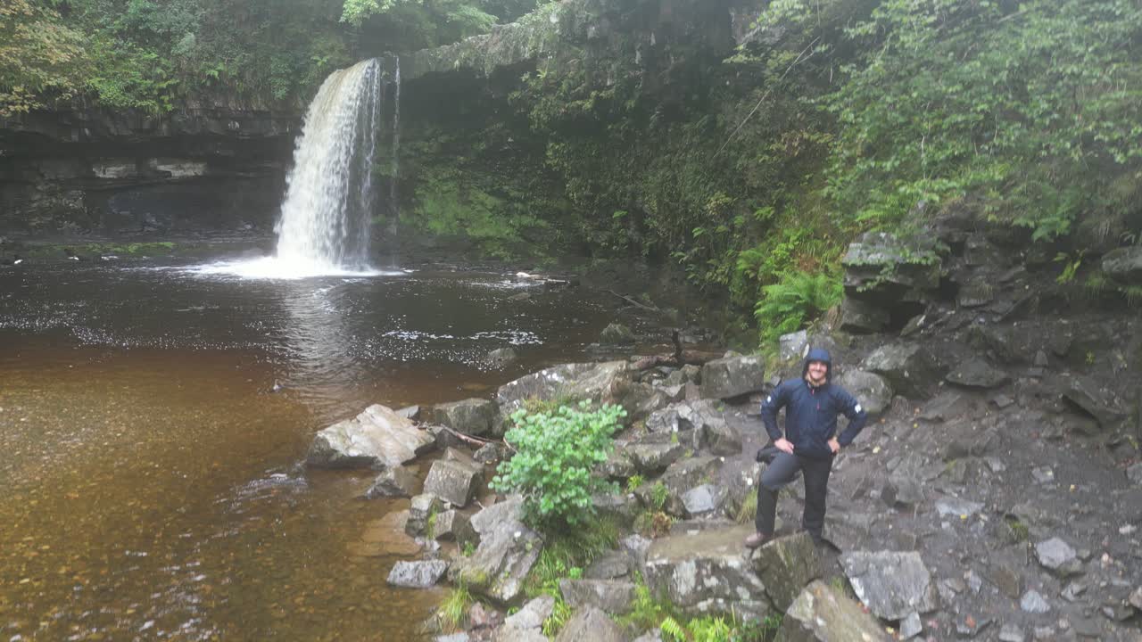 Aerial footage of a man posing by Angel Falls in Brecon Beacons, surrounded by vibrant greenery and rocky terrain on a damp, overcast day. Cascading waterfall adds drama