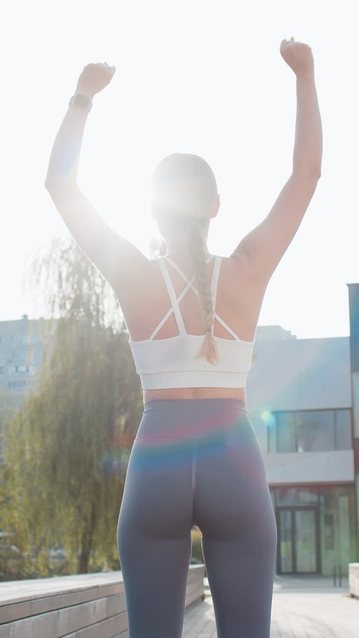 Rear view of young woman runner raising arms celebrating success after jogging in urban city park