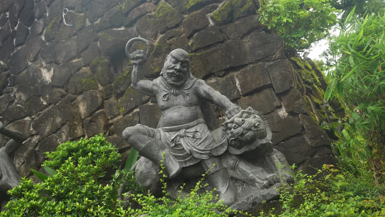 A low static angle holds on the stone sculpture. The muscular figure grips a circular object, subduing a lion-like beast. The fixed frame captures tension, carved detail, and lush natural contrast