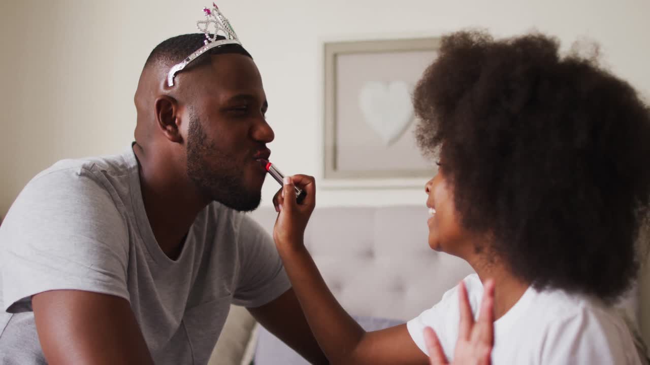 African american girl applying lipstick to her father father wearing crown at home