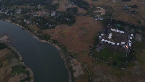 Aerial-view-of-gas-plant-in-the-middle-of-rice-paddy-field-on-the-bank-of-Surma-river-after-sunset,-Bangladesh