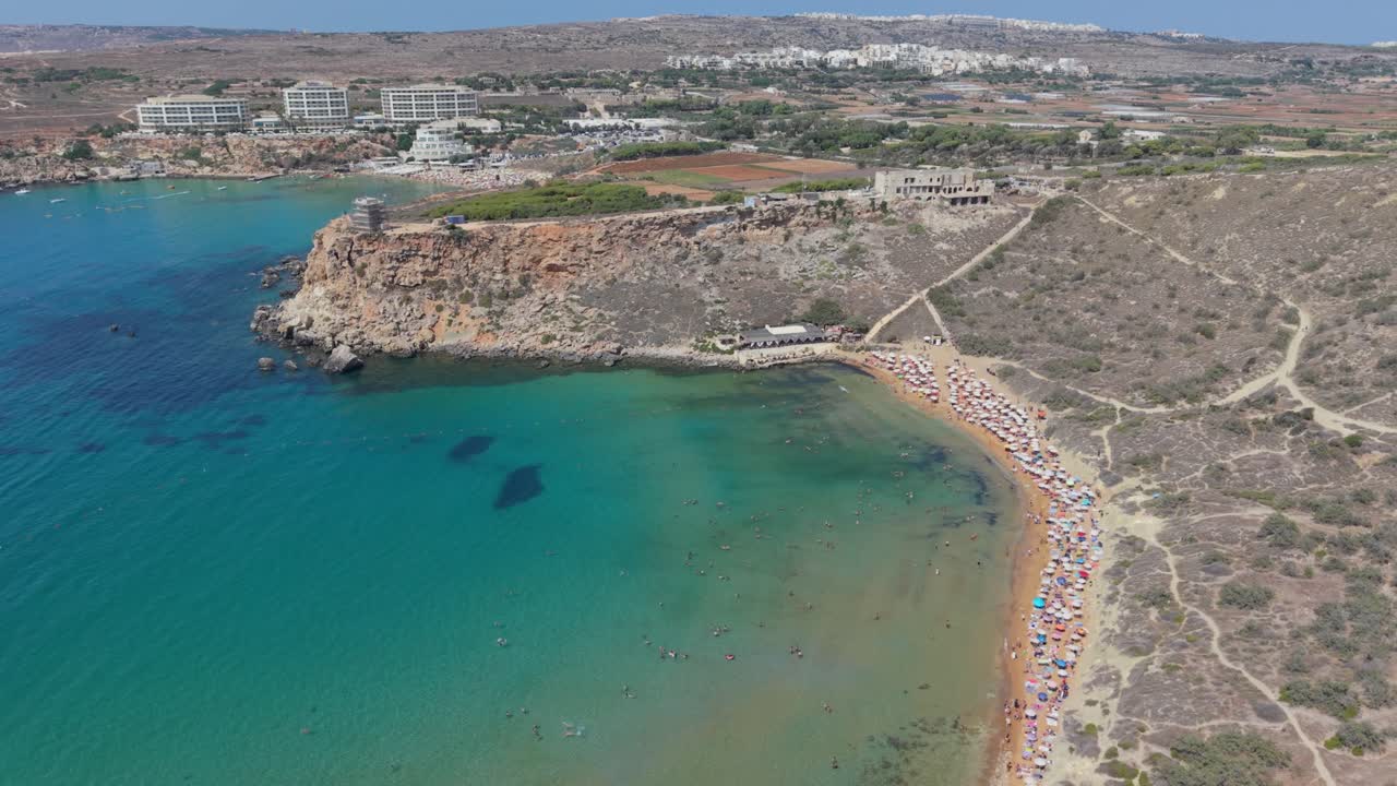 Drone video of a bay with turquoise waters and a sandy beach filled with colorful umbrellas and sunbathers. Swimmers are scattered along the shallow shoreline while rocky cliffs rise in the background