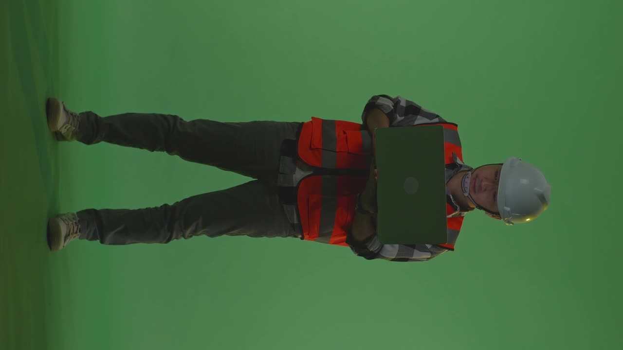 cuerpo lleno de ingeniero masculino asiático con casco de seguridad trabajando en una computadora portátil mientras está de pie en el estudio de fondo de pantalla verde, durante la puesta o el amanecer