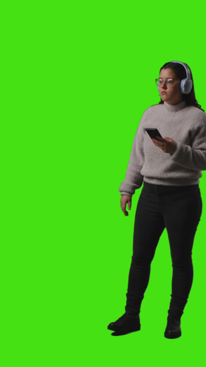 video vertical de larga duración de una mujer con auriculares inalámbricos transmitiendo música desde un teléfono móvil contra una pantalla verde 1