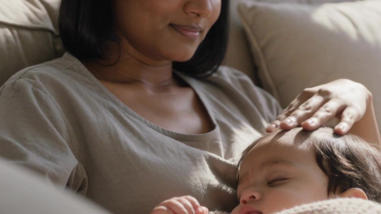 Noticing baby sleeping, mom cradling, stroking baby on couch to soothe with cream blanket