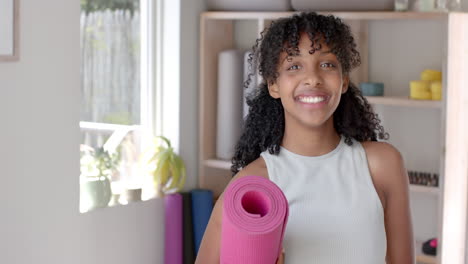 Retrato-De-Una-Feliz-Adolescente-Birracial-De-Fitness-Sosteniendo-Una-Estera-De-Yoga-En-Una-Habitación-Blanca,-Cámara-Lenta