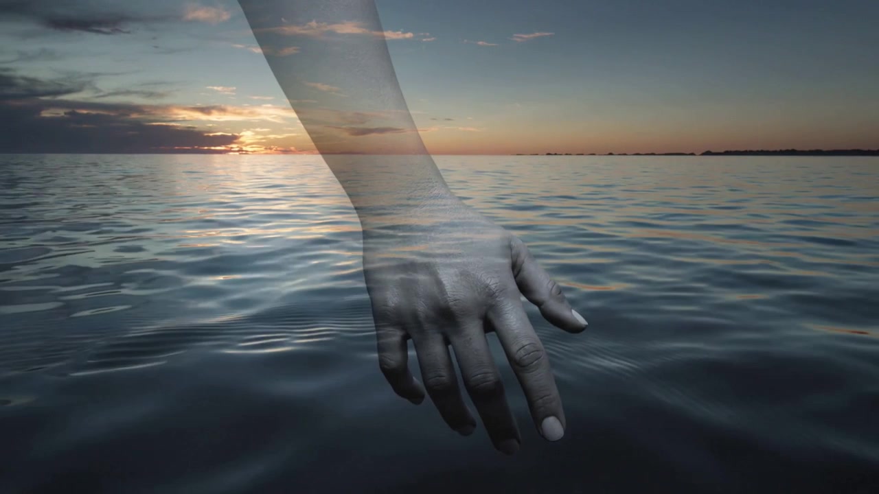 Entering frame, translucent forearm and hand skimming calm sea at sunset, creating ripples