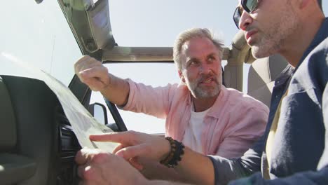 Happy-caucasian-gay-male-couple-sitting-in-car-reading-map-and-pointing-on-sunny-day-at-the-beach