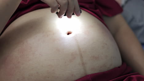 an expectant mother is holding a tiny flashlight and moving it around her swollen belly, showing stretched marks and some scratches on her skin