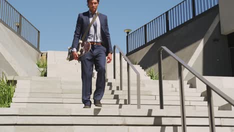 Caucasian-businessman-walking-downstairs,-holding-skateboard-on-sunny-day