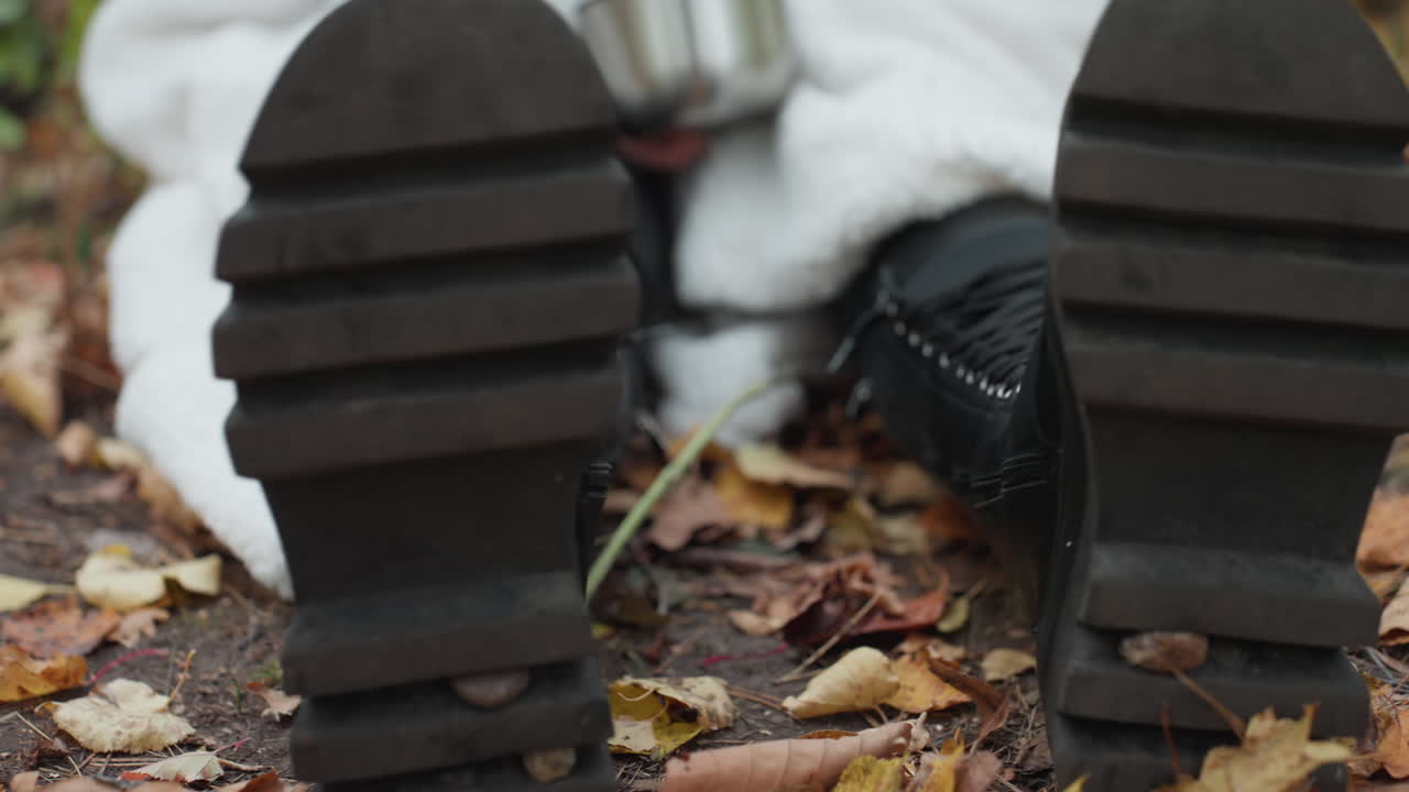 close up sepatu bot kulit di atas daun musim gugur, wanita berbalut selimut memegang cangkir logam, tampilan sudut rendah menyoroti tapak berat dan sol yang usang, suasana berpasir dengan corak warna tanah dan cahaya mendung yang lembut