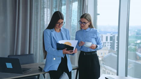two female coworkers stand near window at office. office employees discussing work issues looking at documents. office indoors backdrop.