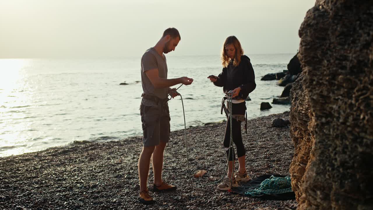 A blonde girl with curly hair in black clothes and a brunette guy in a gray T-shirt and gray pants tie special knots on their belay using special carabiners in order to start rock climbing on a rocky seashore near the rocks