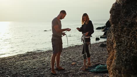 A-blonde-girl-with-curly-hair-in-black-clothes-and-a-brunette-guy-in-a-gray-T-shirt-and-gray-pants-tie-special-knots-on-their-belay-using-special-carabiners-in-order-to-start-rock-climbing-on-a-rocky-seashore-near-the-rocks