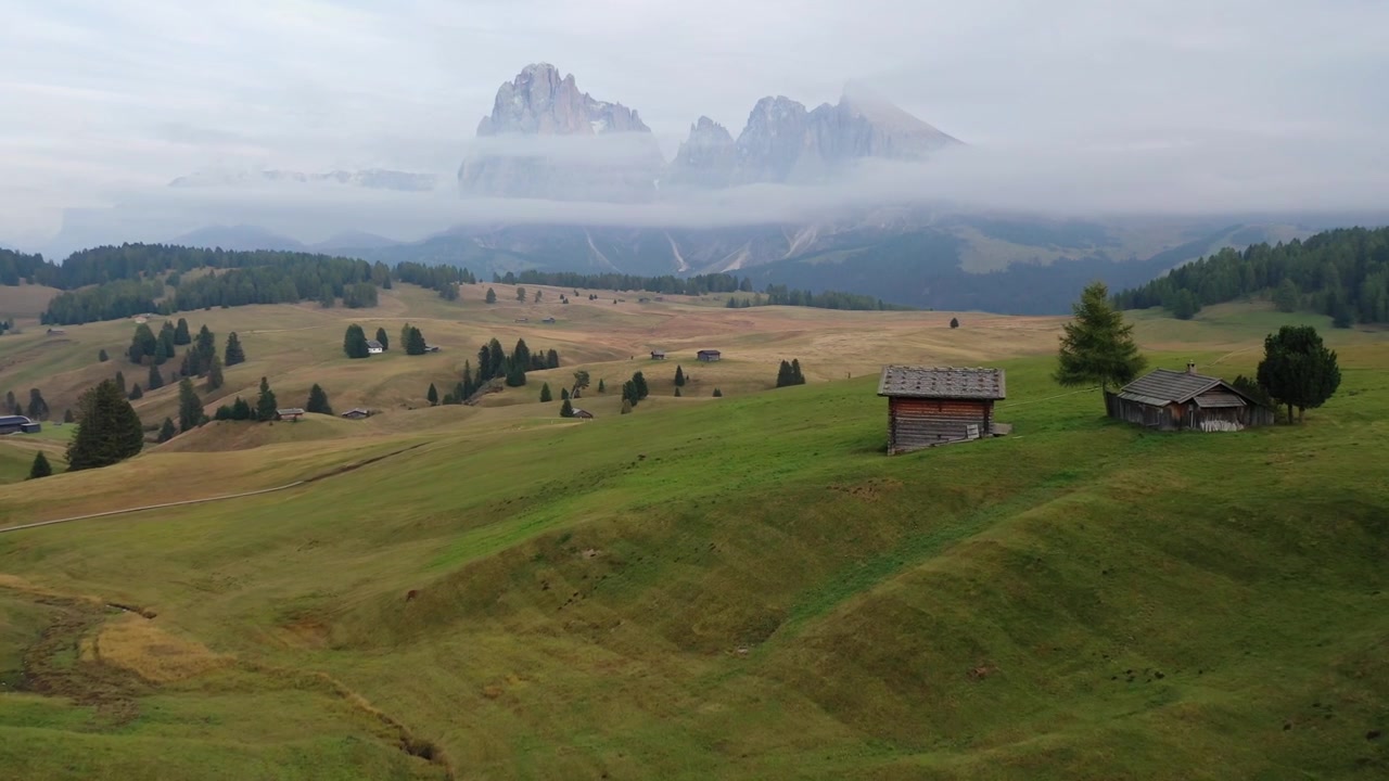 Carrellata da dietro di una donna che cammina tra le dolci colline verdi dell'Alpe di Siusi. Presenta capanne di legno tradizionali con le imponenti montagne del Sassolungo coperte di nuvole sullo sfondo.