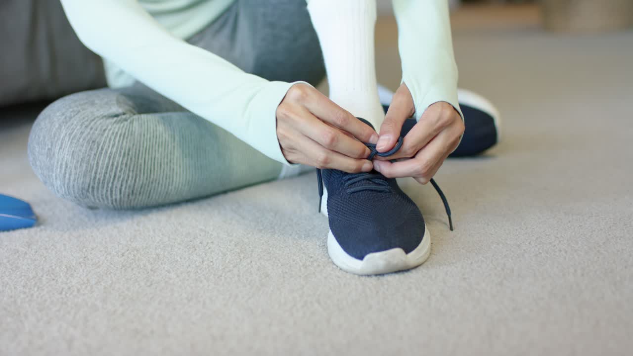Donna afroamericana seduta sul tappeto, che allaccia i lacci delle scarpe da ginnastica blu scuro, preparandosi per l'esercizio fisico