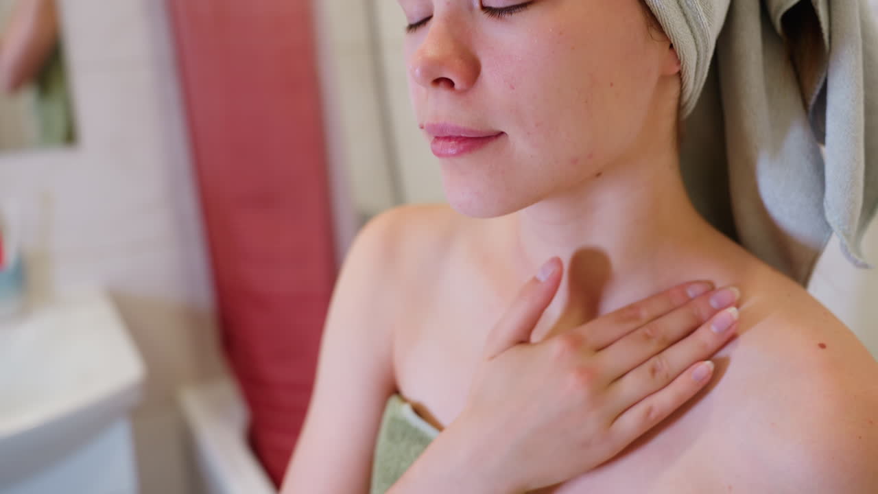 Close-up view of woman applying cream on neck and chest area, she is focused and gentle, moisturizing her skin while wrapped in towel, taking care of her body after shower
