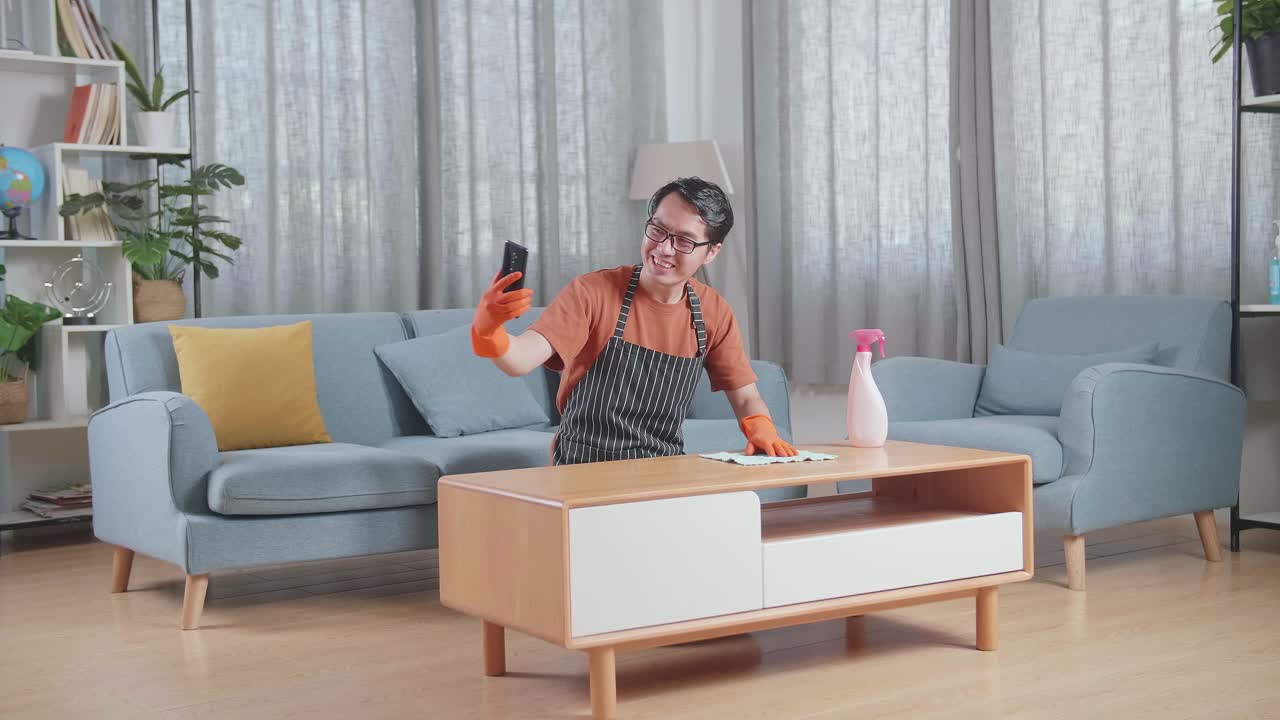 집에서 스프레이로 테이블을 청소하는 동안 스마트폰에서 비디오 통화를 하는 아프론을 입은 아시아 남성 집주인