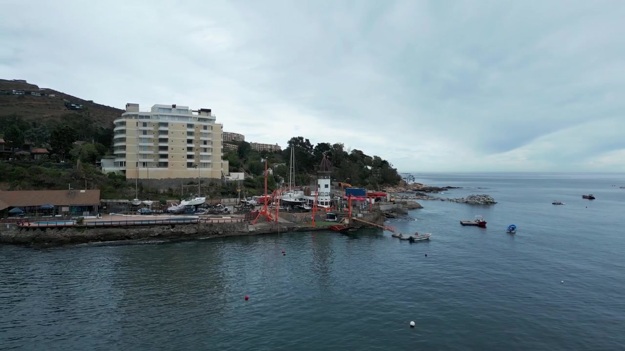 Volo circolare sopra un cantiere navale costiero con gru arancioni e barche ancorate, accanto a una moderna torre di appartamenti su un promontorio roccioso sotto un cielo coperto.