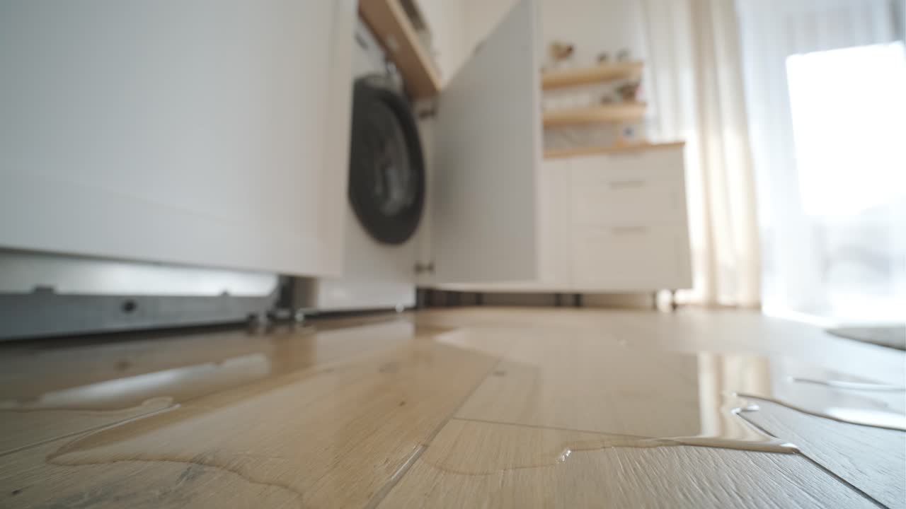 un charco de agua en el suelo de madera de un lavador de platos abierto en una cocina iluminada por el sol, señalando un accidente doméstico