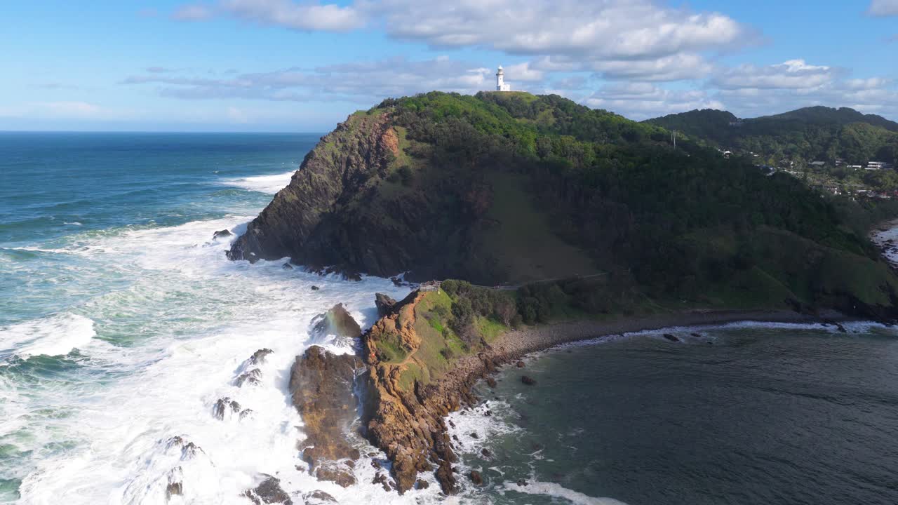 Aerial footage of Byron Bay's rugged coastline and iconic lighthouse under clear skies, showcasing dynamic ocean waves and lush greenery