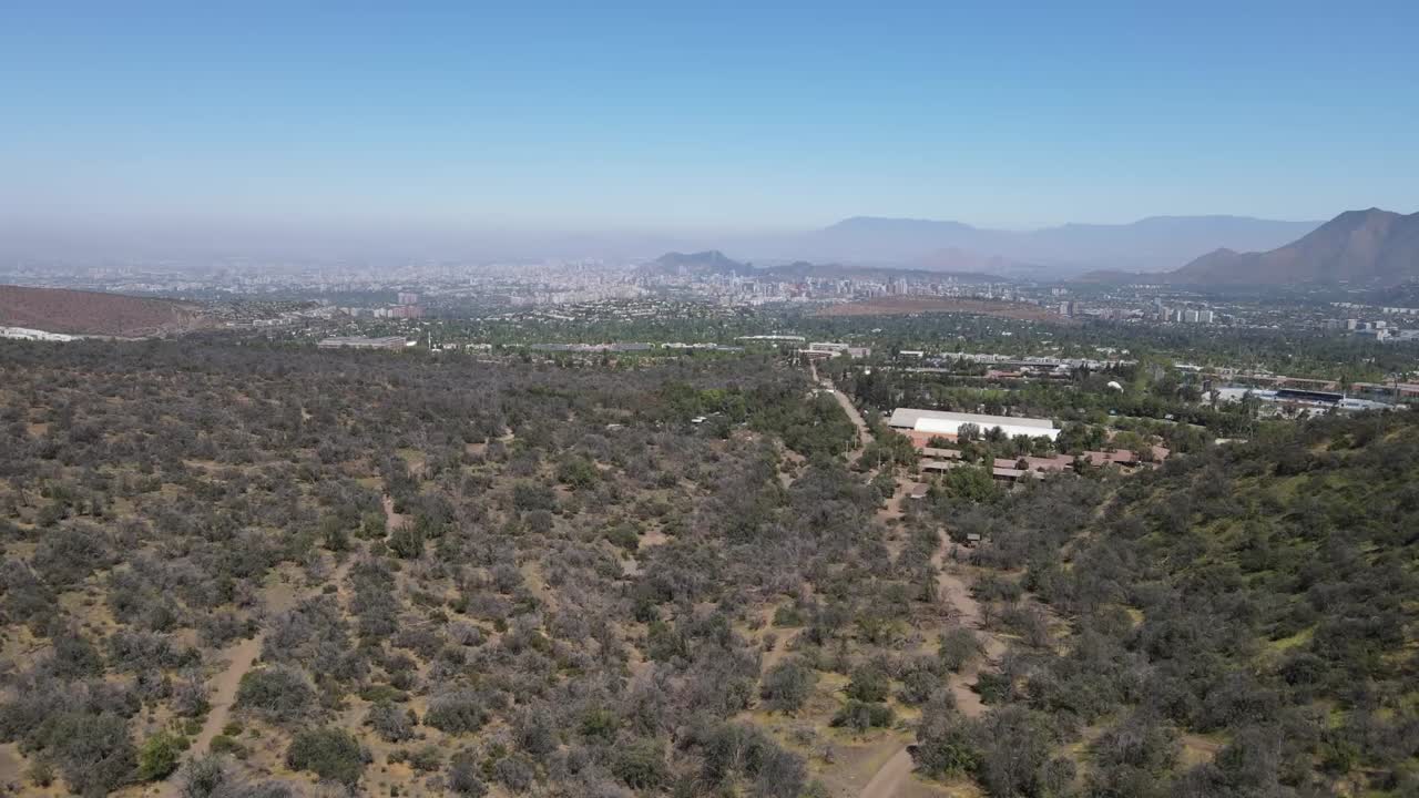 "Dry bushes and hills dominate the foreground, while santiago's skyline gradually appears, veiled in a slight haze under a clear, sunny sky