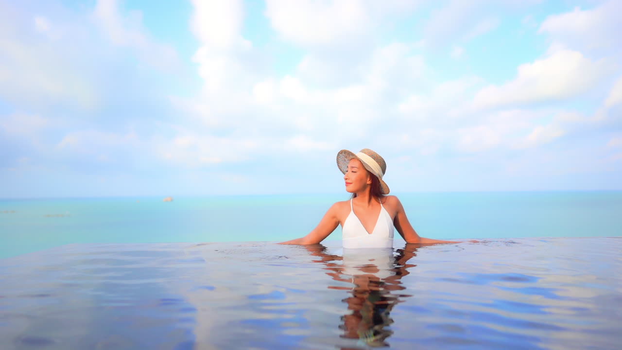 화창한 날 슬로우 모션으로 태국의 청록색 바다 배경에 있는 인피니티 풀 가장자리에 팔을 기대고 있는 흰색 수영복에 맞는 여성 모델
