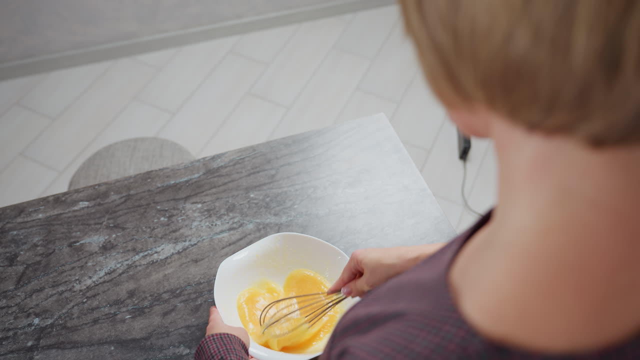 vista trasera de una mujer de cabello dorado con vestido a cuadros mientras agita el huevo en un cuenco, la iluminación suave destaca su postura y enfoque, con una encimera borrosa y configuración de cocina