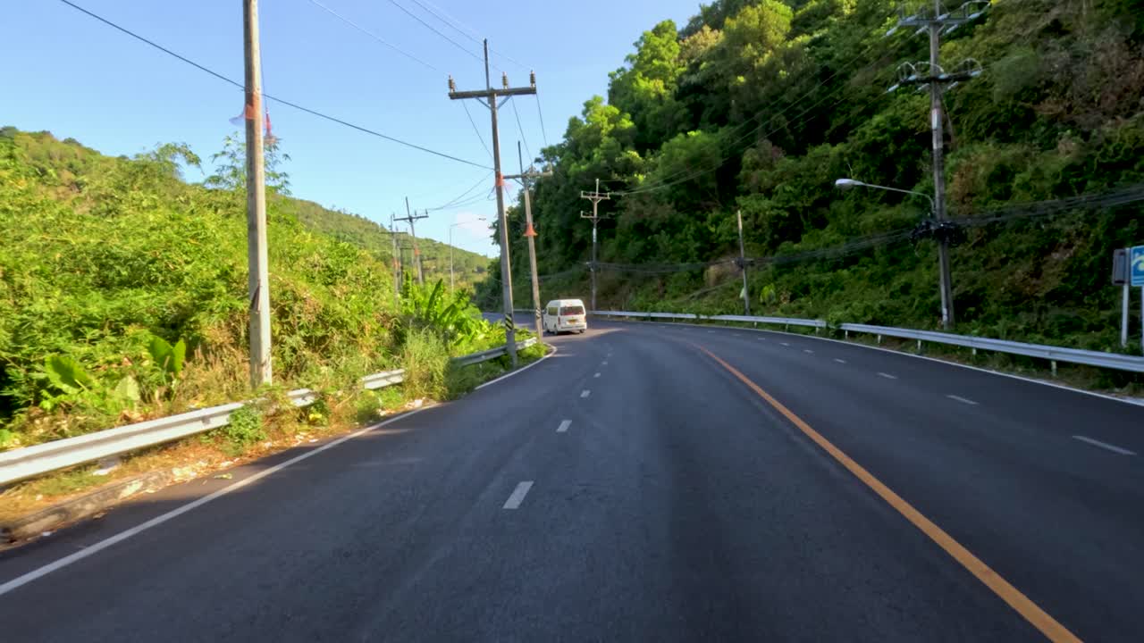 Vehicles navigate a curving, sunlit road through lush tropical hills in Phuket, Thailand