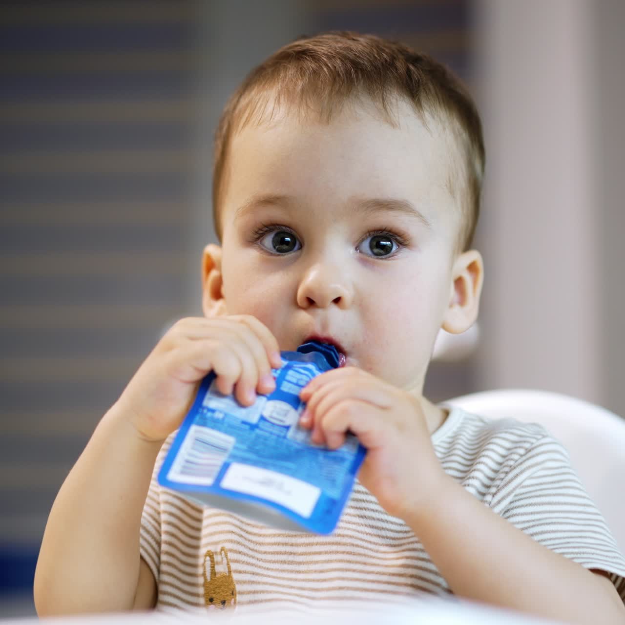 Beautiful toddler consuming factory produced fruit puree. Baby boy eats organic food from doy pack. Close up. Blurred backdrop