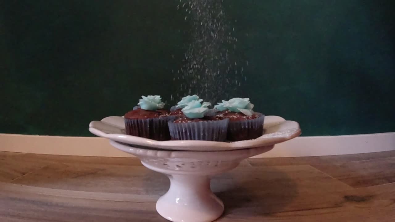 Chocolate cupcake muffins with a blue sugarpaste rose on top being covered in sugar powder as if it's snowing. Steady slow motion camera shot.