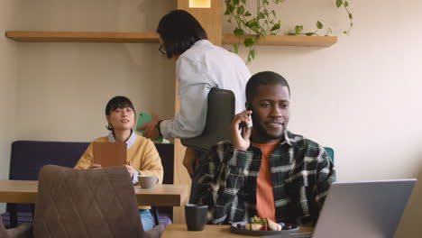 Hombre-Hablando-Por-Teléfono-Celular-Mientras-Trabaja-Con-Una-Computadora-Portátil-En-El-Café-Mientras-El-Camarero-Toma-El-Pedido-De-La-Mujer-Detrás-De-él