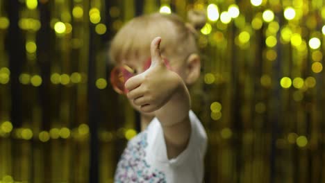 Child-show-thumbs-up,-smiling,-looking-at-camera.-Girl-posing-on-background-with-foil-golden-curtain