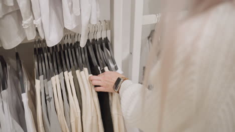 high angle view shows woman with pastel hair browsing rack, sliding hangers with manicured hand wearing gold chain watch, assessing neutral cotton tops beneath white shirts inside bright boutique