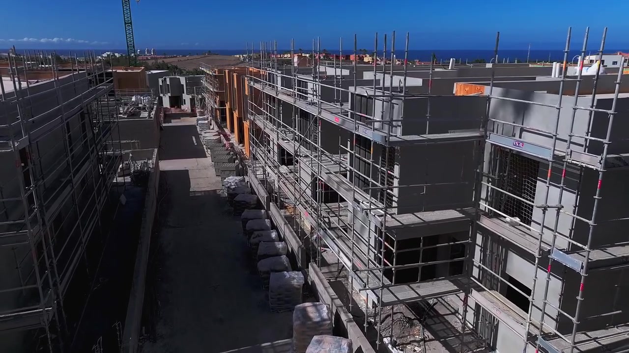 Drone view through an active residential construction site, showing partially completed modern apartment buildings, scaffolding, cranes and ongoing facade and interior works near the coast