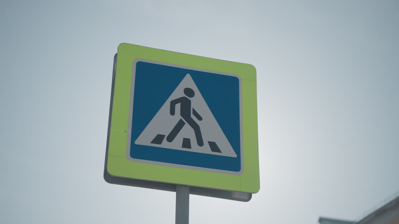 close view pedestrian crossing sign on pole under bright winter sun, blue triangle symbol against pale sky, urban street safety marker guiding walkers and drivers through cold season traffic, crisp light, calm weather