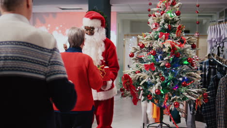 Employee-wearing-Santa-Claus-suit-holding-raffle-contest-in-Christmas-decorated-shopping-mall-clothing-shop-during-winter-holiday-season,-inviting-elderly-couple-to-participate