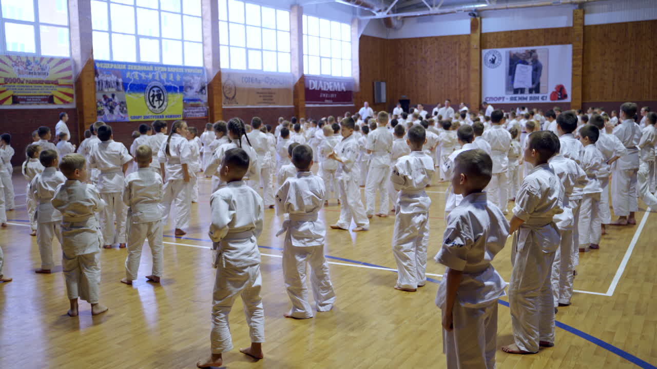 Spacious gym crowded with numerous young sportsmen standing in rows. Teenage karate trainees applauding to coaches.