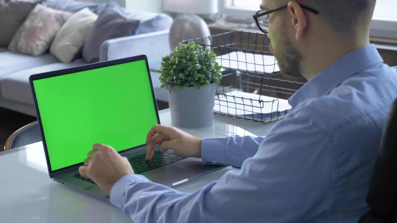 hombre usa una computadora portátil con pantalla de maqueta verde mientras está sentado en el escritorio en su acogedora sala de estar