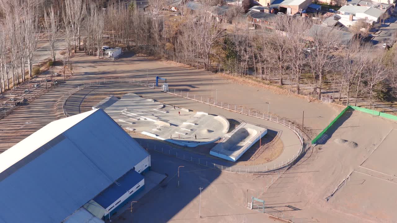 Aerial orbiting drone shot of a skate park next to a large sports hall in Malargüe, Mendoza, Argentina, with bare trees and long shadows suggesting late autumn
