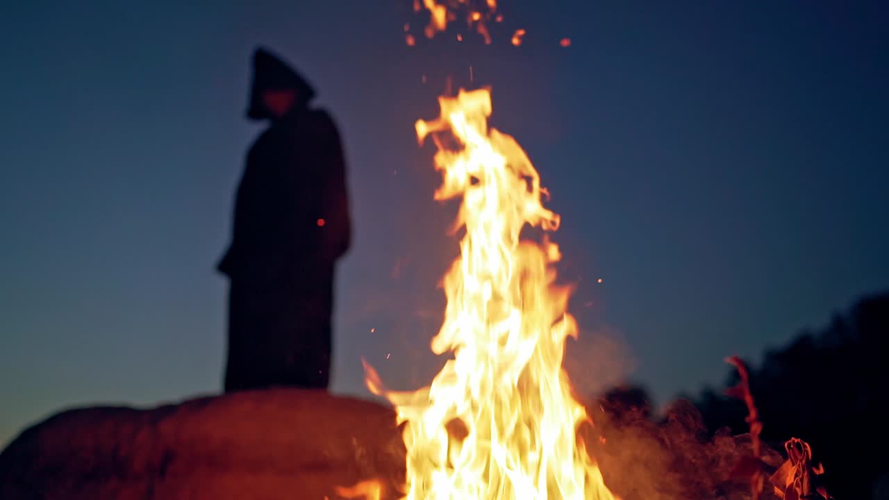 Huge flame and some smoke on the background of black figure outdoors. Silhouette of death on the hill at night. Vivid flame and spooky person.