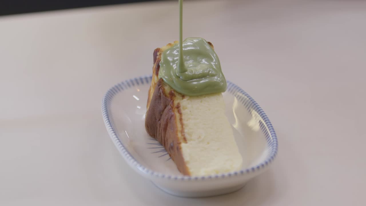 A visually appealing, close-up shot of thick green cream sauce (likely matcha or pistachio) being poured over a slice of Burnt Basque Cheesecake served on a small ceramic plate
