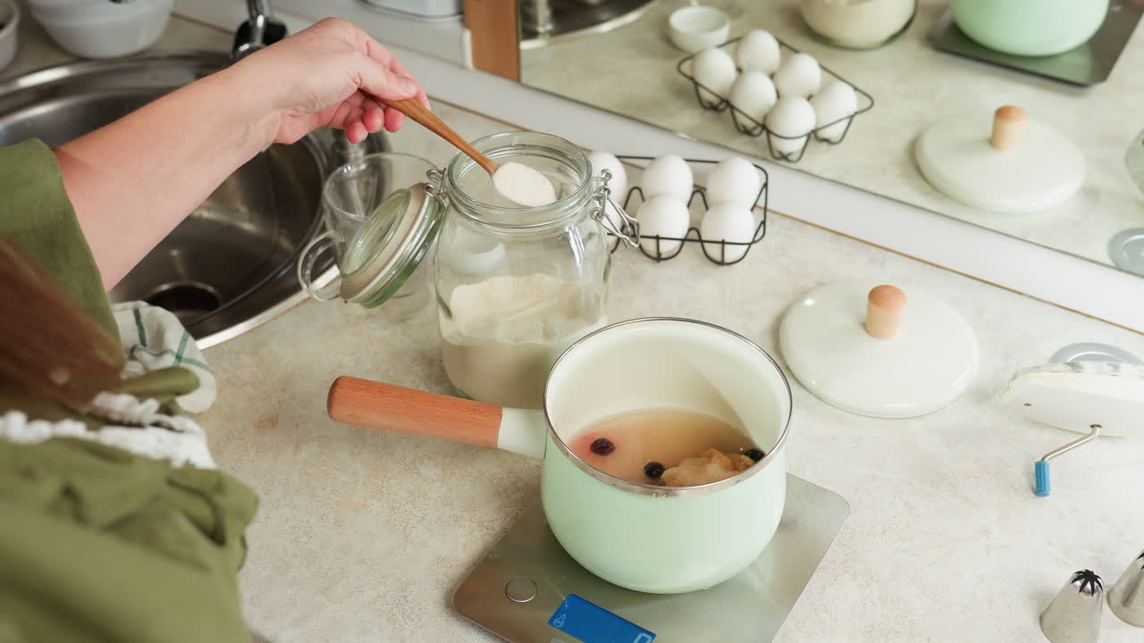 Chef scooping flour from transparent jar into pastel green cooking pot with fruit mixture placed on digital kitchen scale, surrounded by eggs, tools, and utensils in bright kitchen setup