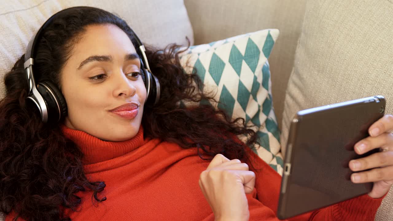 Happy woman lying on sofa using tablet enjoying music on her headphones 4K 4k