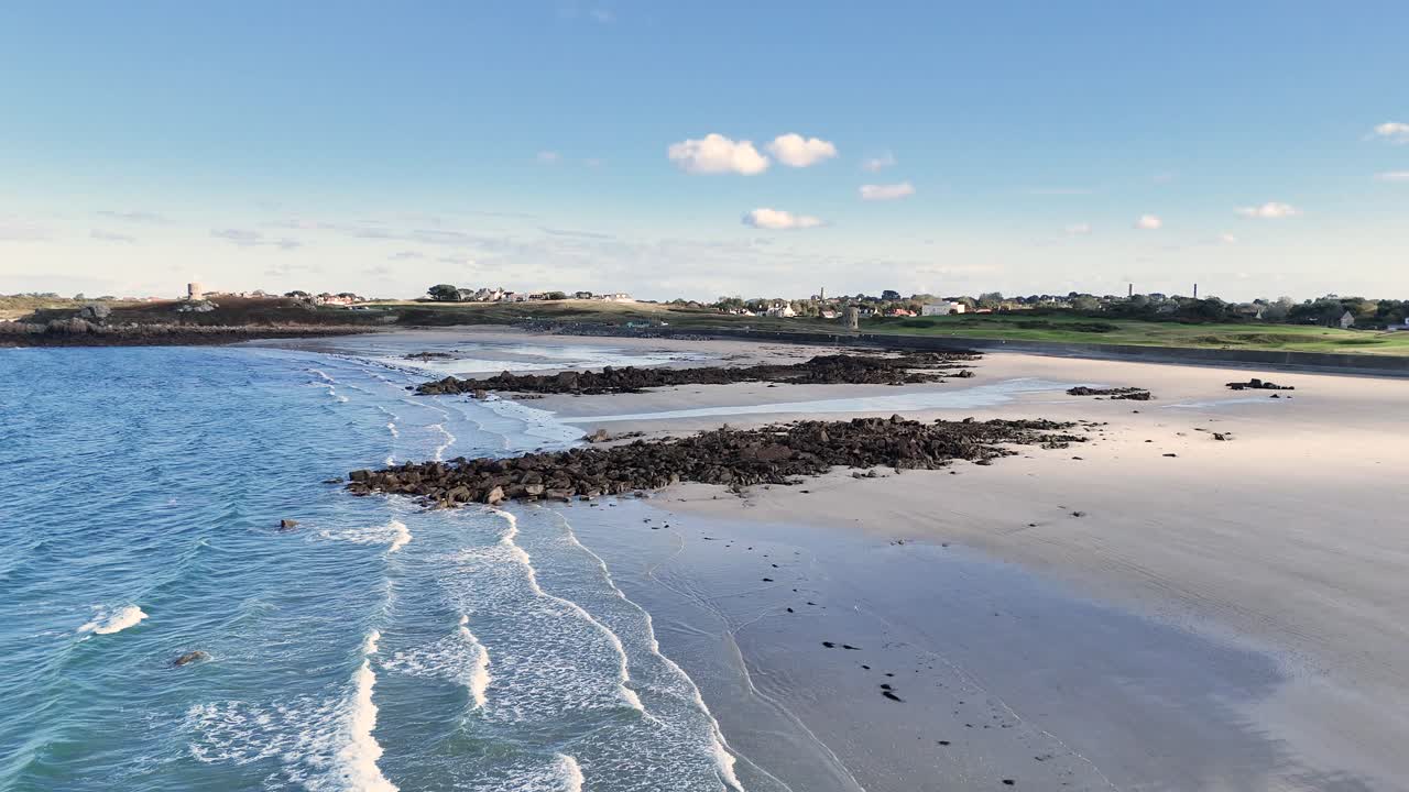 Lancresse Bay Guernsey flight over shoreline of golden sandy beach with rocky outcrops on sunny day with calm lapping waves and views over Lancresse Common