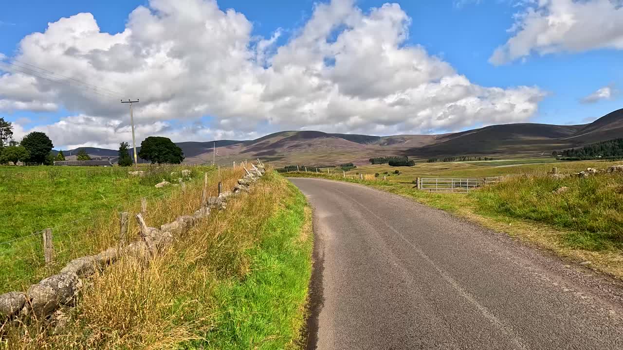 A vehicle travels a winding single-lane road bordered by stone walls and green fields under bright daylight, with rolling hills and scattered clouds visible