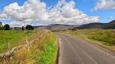 a vehicle travels a winding single-lane road bordered by stone walls and green fields under bright daylight, with rolling hills and scattered clouds visible