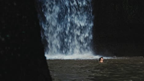 Tall-waterfall-in-slow-motion-in-Bali,-Indonesia-and-man-swimming-in-water